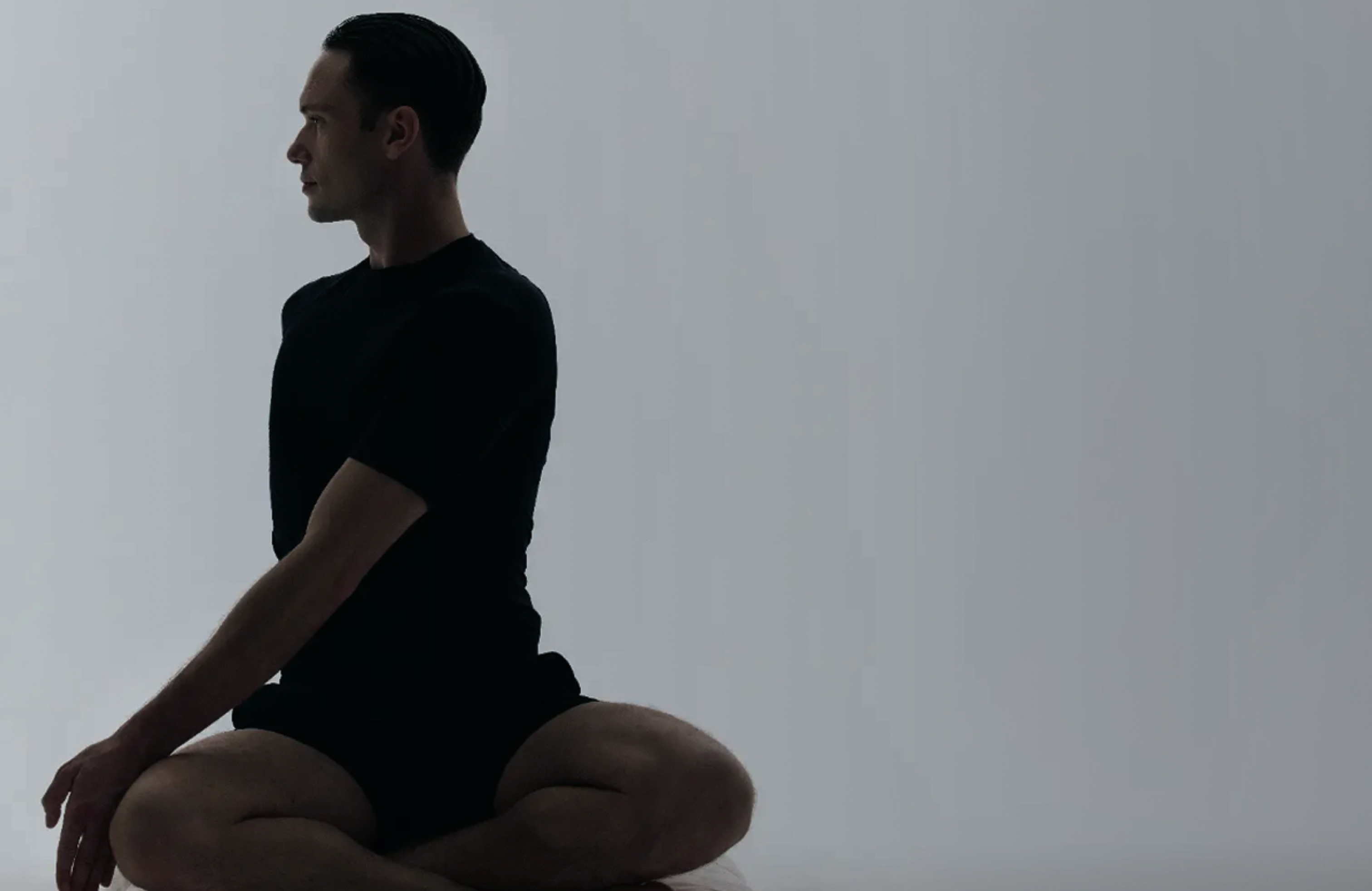 A man sitting cross-legged in profile against a soft grey studio backdrop